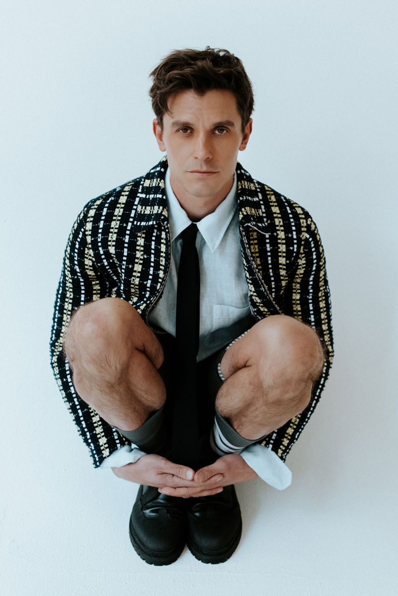 Antoni Porowski crouching in a patterned tweed jacket, collared shirt, and black tie against a white studio background.