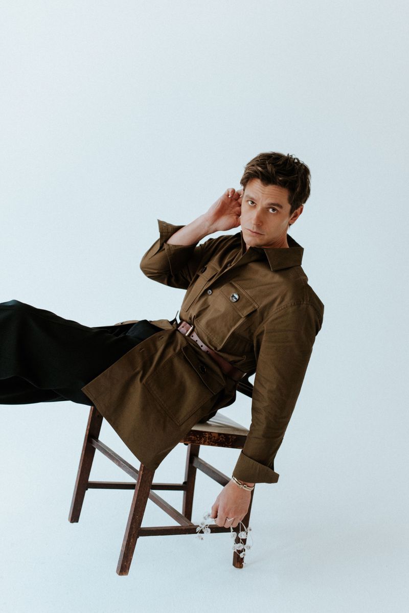 Antoni Porowski in an olive green belted jacket and black trousers leaning on a wooden chair against a white seamless studio backdrop.