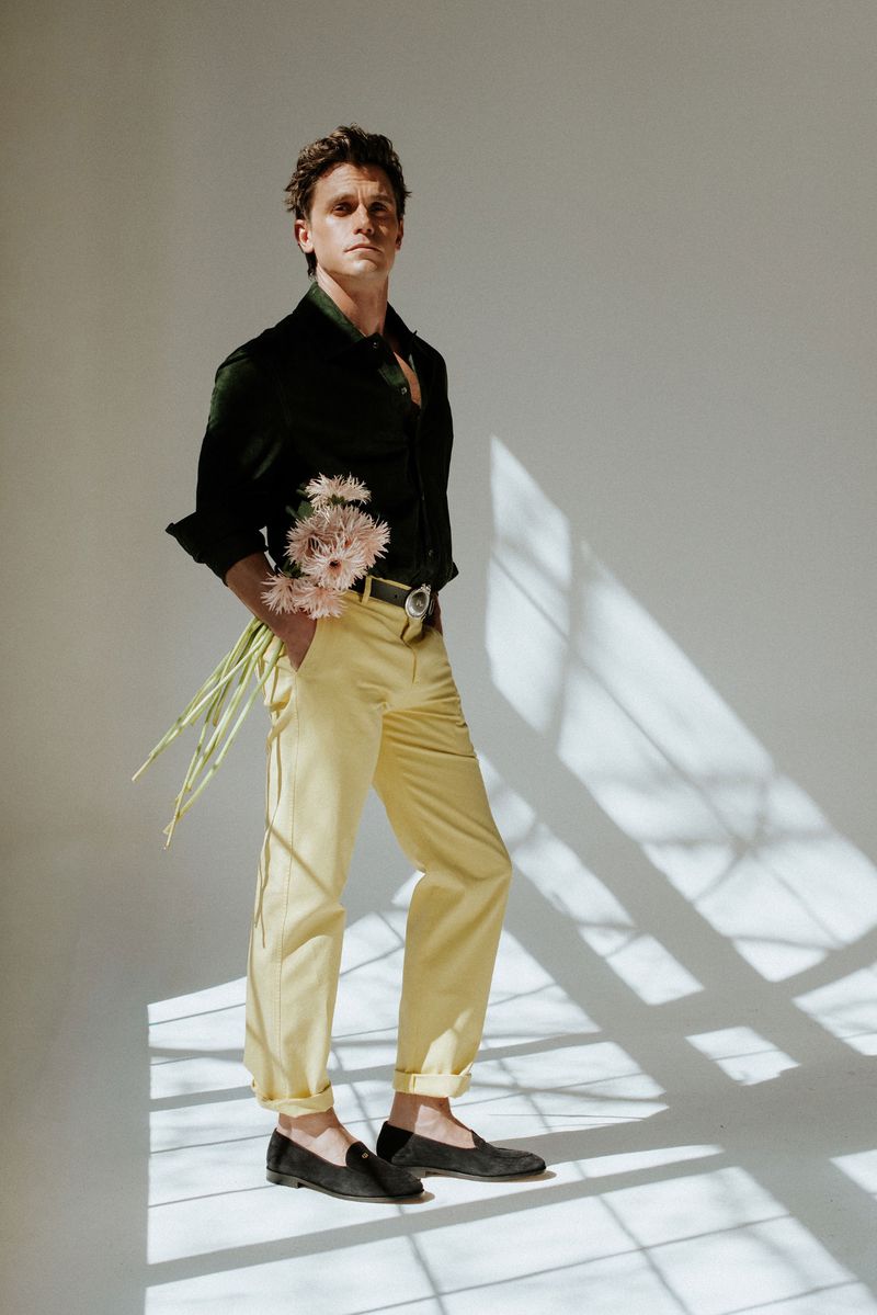 Antoni Porowski poses holding pink flowers in a black shirt and yellow trousers in a sunlit white studio.