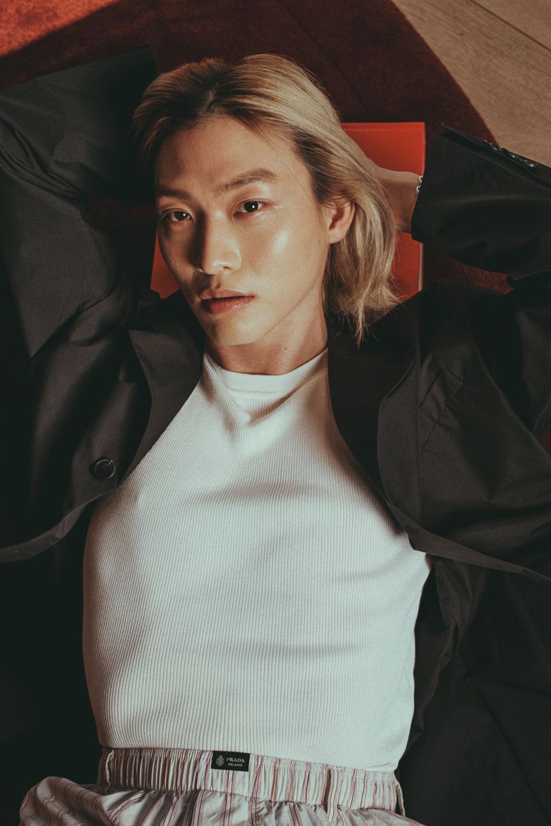 High-angle shot of a blonde East Asian man lying down wearing a white ribbed tank top and black coat.