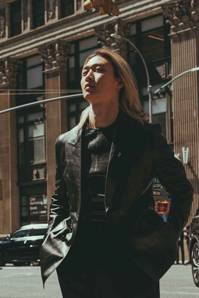 An East Asian man with blonde hair poses on a city street wearing a black leather blazer in direct sunlight.