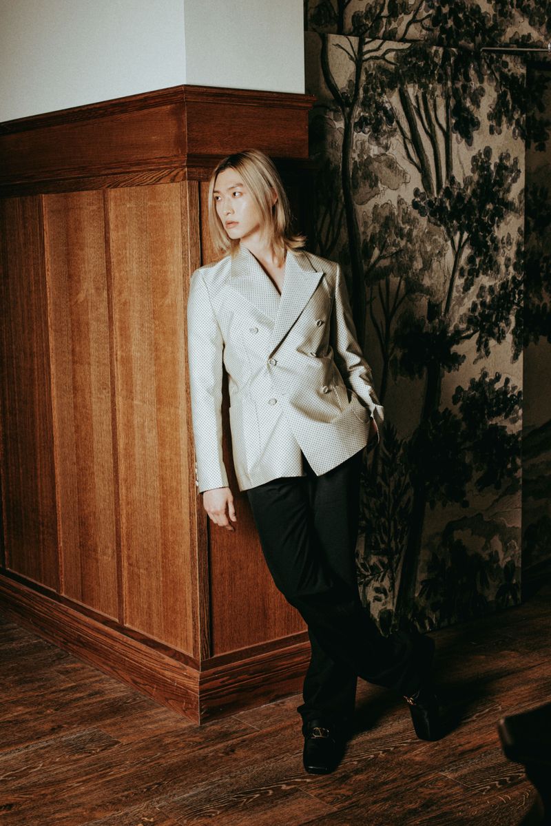 Model with blonde hair wearing an off-white double-breasted blazer and black trousers, leaning against a rich wood-paneled wall.