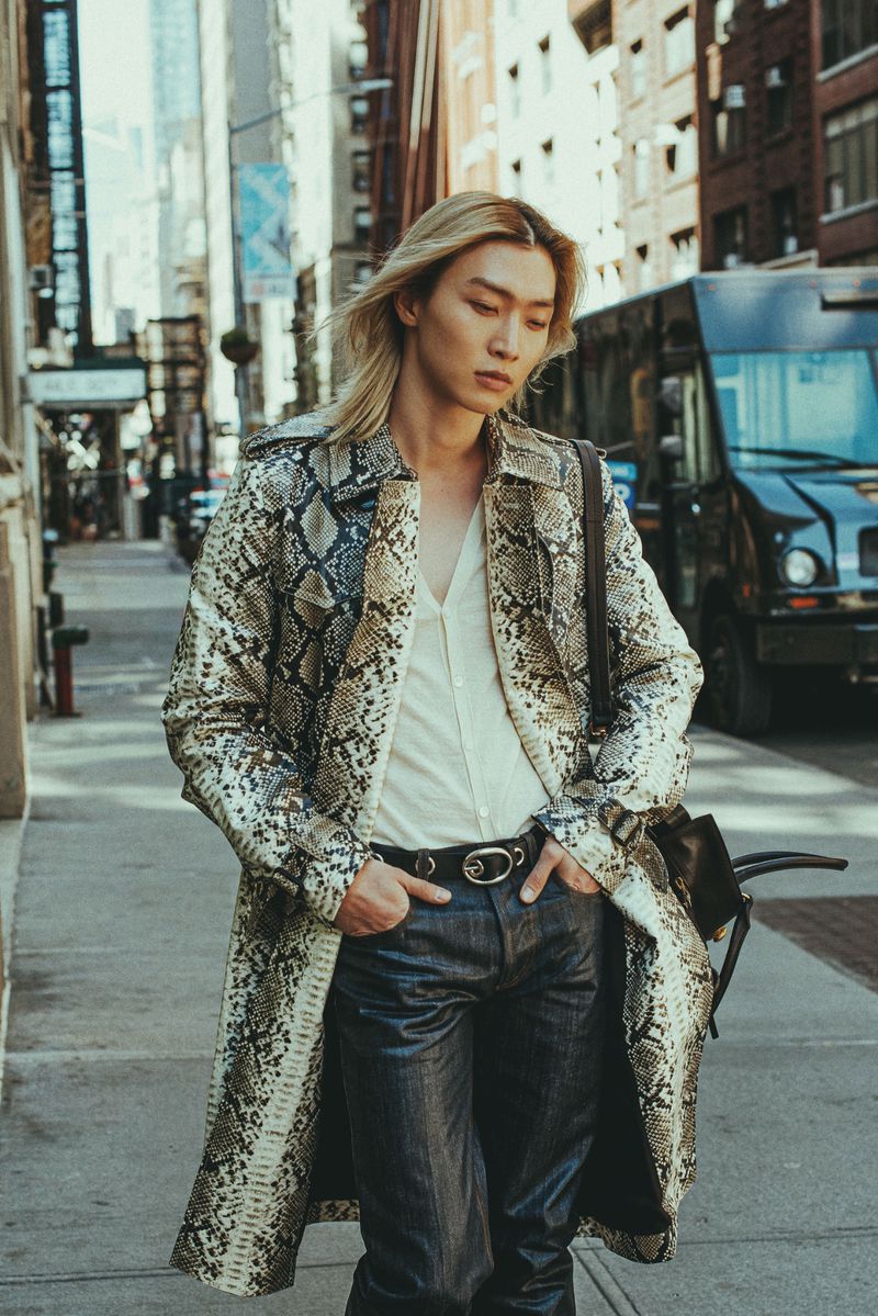 East Asian man with blonde hair wearing a snakeskin print trench coat and dark jeans on an urban city sidewalk.