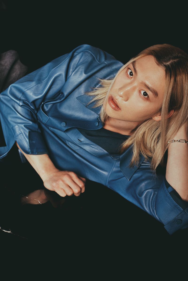 East Asian model with blonde hair reclining in a blue leather shirt against a black background.