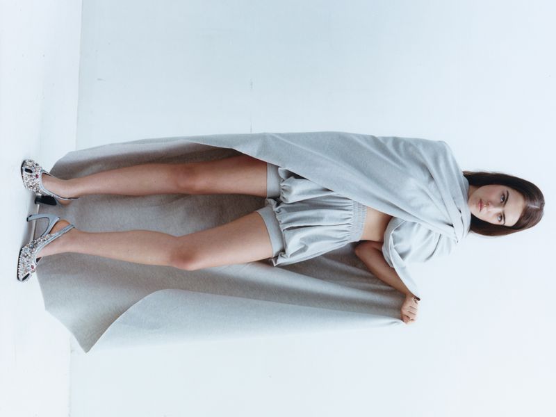Model Stella Jones draped in light grey fabric with matching shorts and embellished slingback heels against a white studio background.