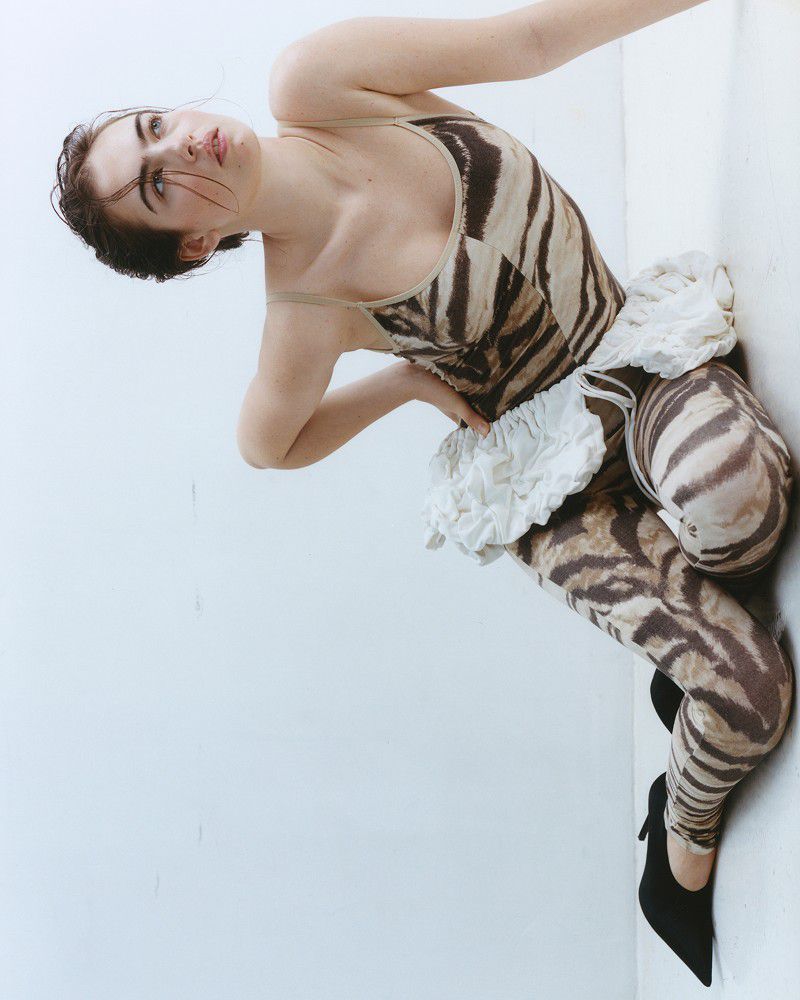 Stella Jones in a brown tiger-print jumpsuit and black heels posing against a white studio background.