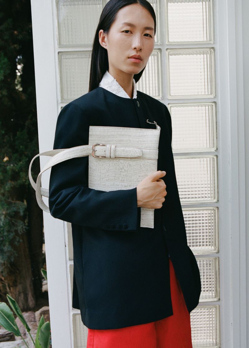 A fashionable Asian woman with long dark hair poses with a shoulder bag in front of a window. She wears a dark blazer and red pants.