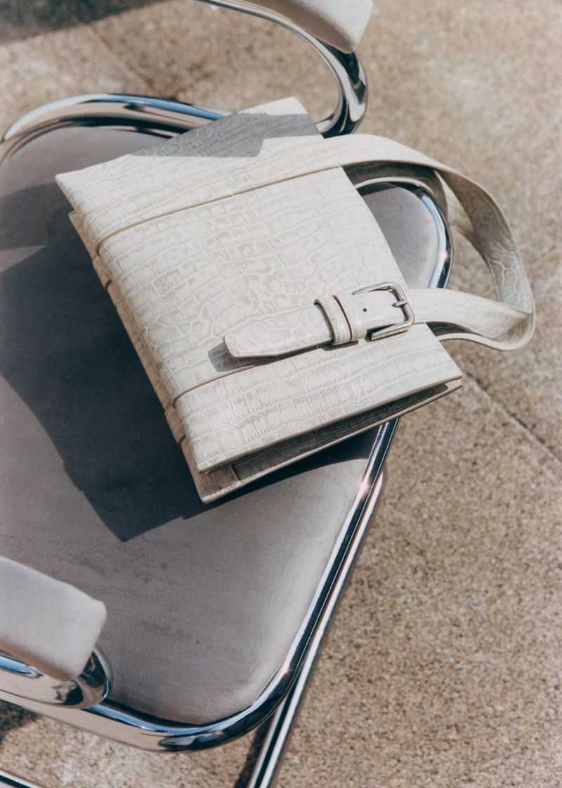 Close-up of a textured off-white bag with a buckle strap resting on a gray and chrome chair with a concrete floor in the background.