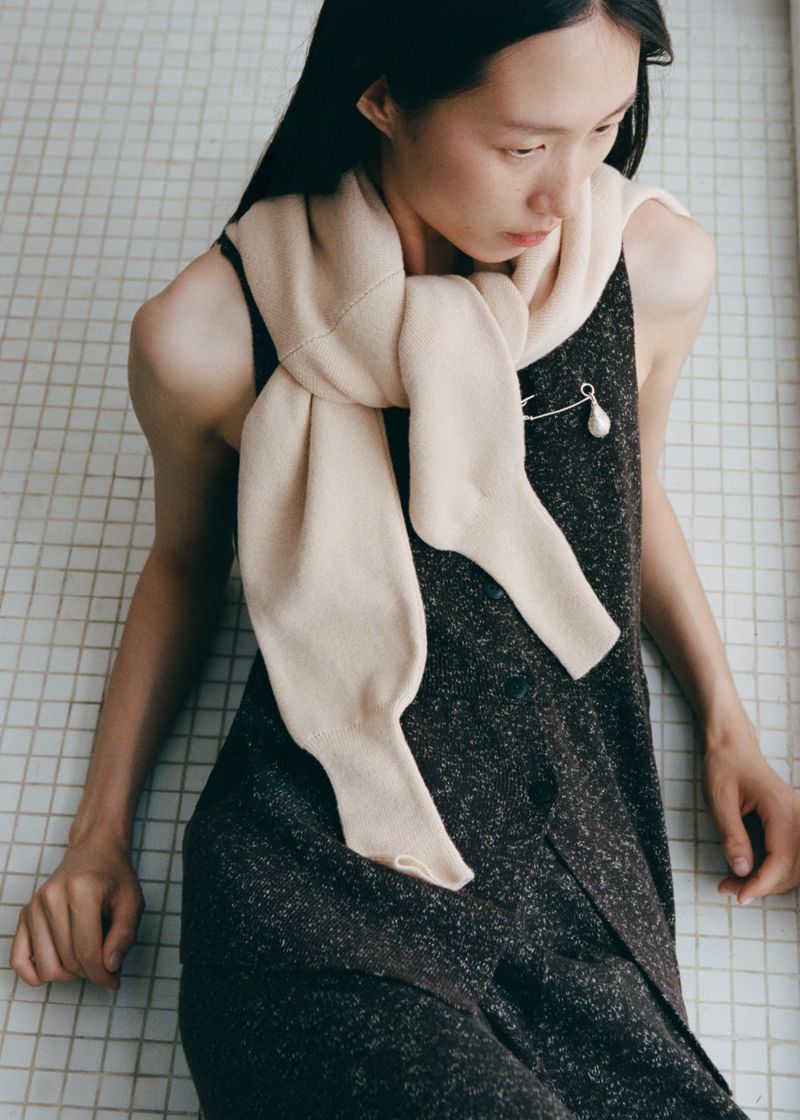 A woman poses in a sleeveless dress with an ivory-colored knit scarf draped over her shoulders in a neutral-toned indoor setting.
