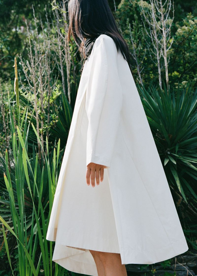 Fashion shot of a brunette woman in a white coat standing in a lush green garden. Elegant, full length coat, natural light.
