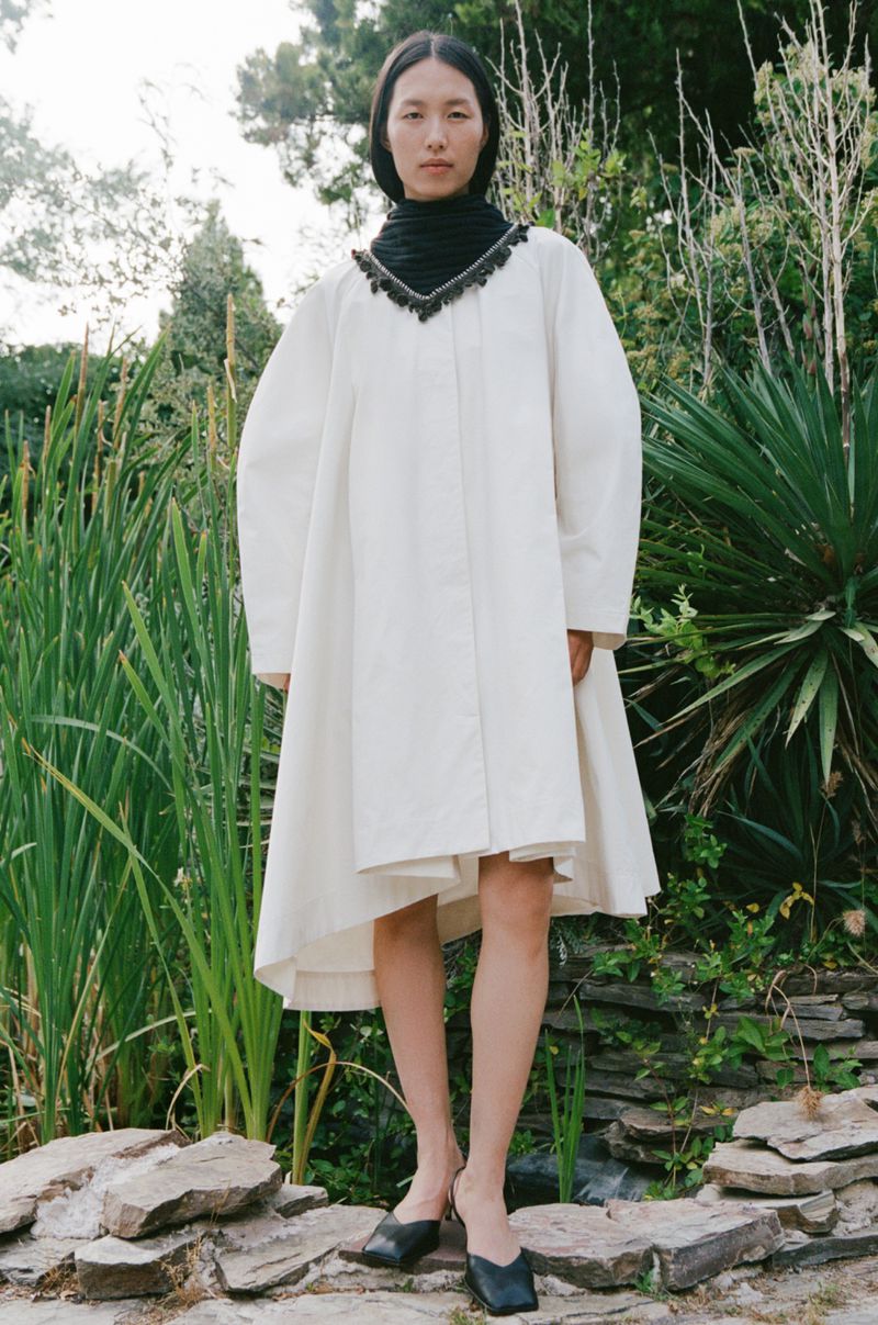An Asian woman poses in a flowing white dress, accented by a black collar with fringe, in a lush outdoor setting with greenery and stone.
