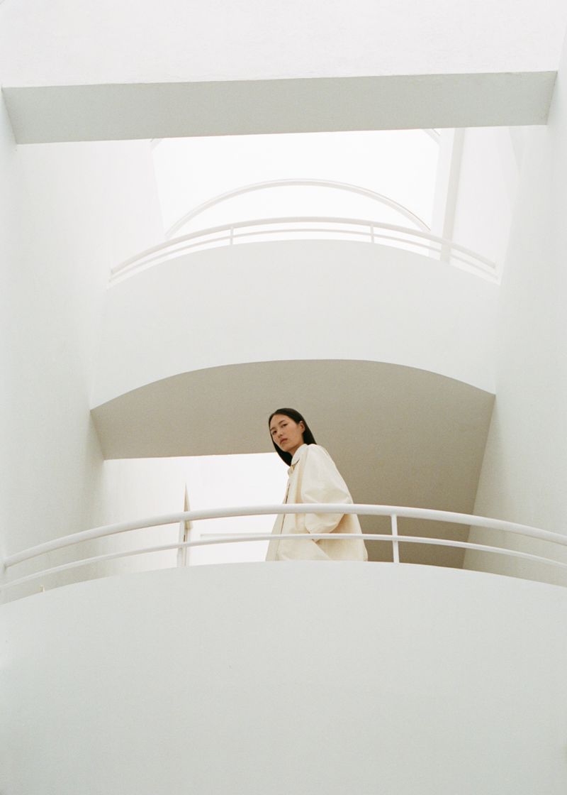 Indoor shot of a young adult woman in a cream-colored coat, standing on a balcony with curved white walls and a high ceiling.
