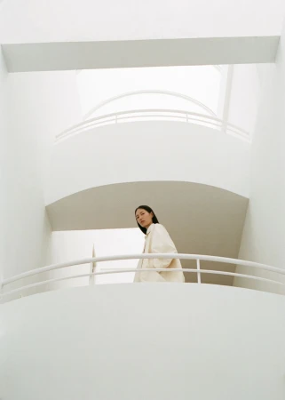 Indoor shot of a young adult woman in a cream-colored coat, standing on a balcony with curved white walls and a high ceiling.