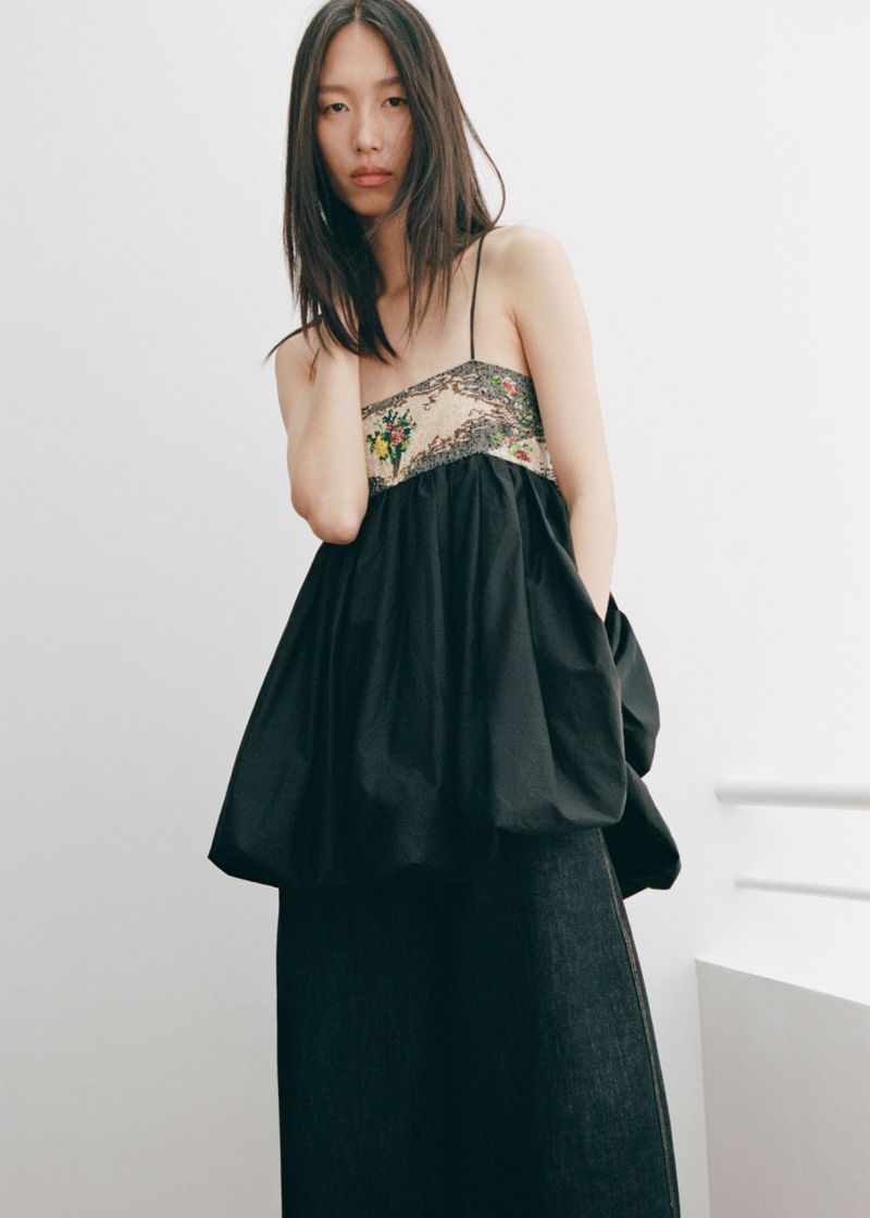 An Asian woman wears a sleeveless top with a floral bodice and flowing black skirt, posing in front of a plain white wall.