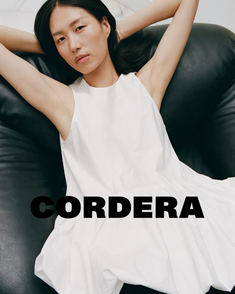 A relaxed young woman in a white dress reclines on a black leather beanbag chair in a fashion-forward editorial setting.