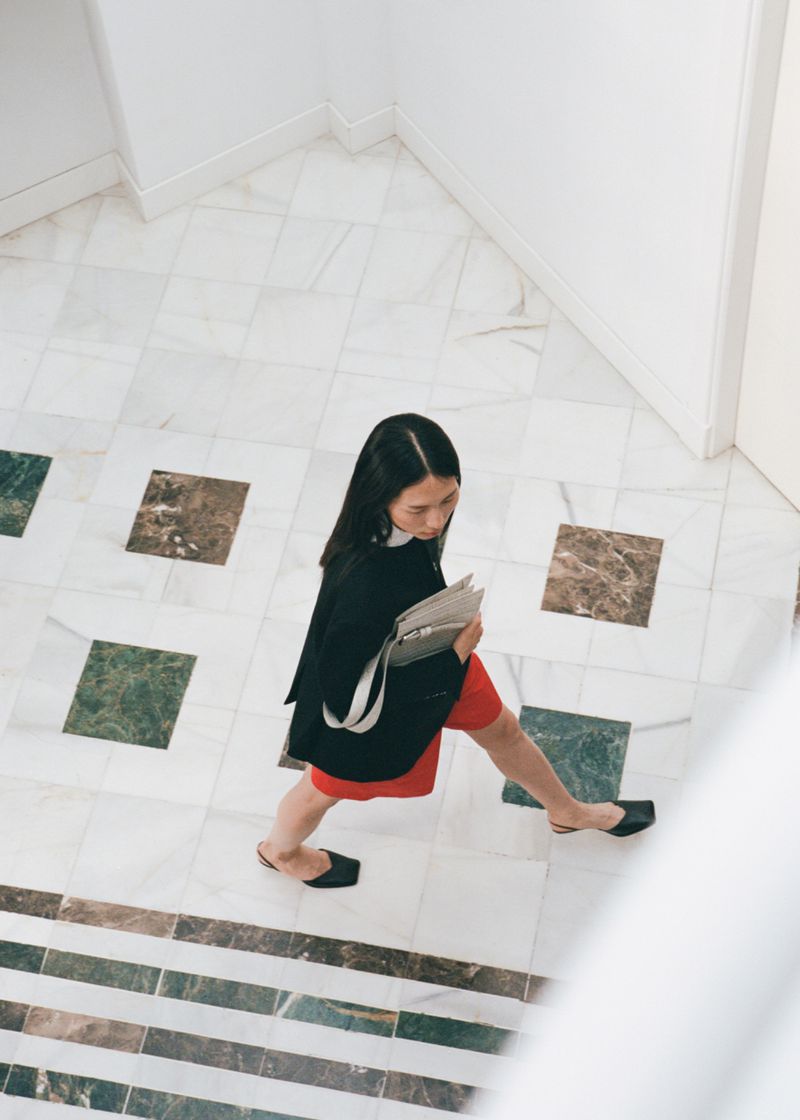 Overhead view of a woman in a black jacket and red skirt walking on a marble tile floor while holding a light-colored bag or document.