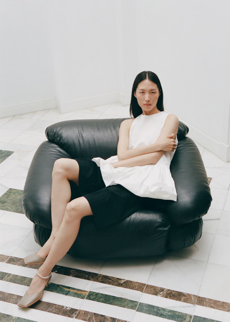 A young Asian woman in a sleeveless white top and black shorts poses on a black leather chair in a bright indoor setting.