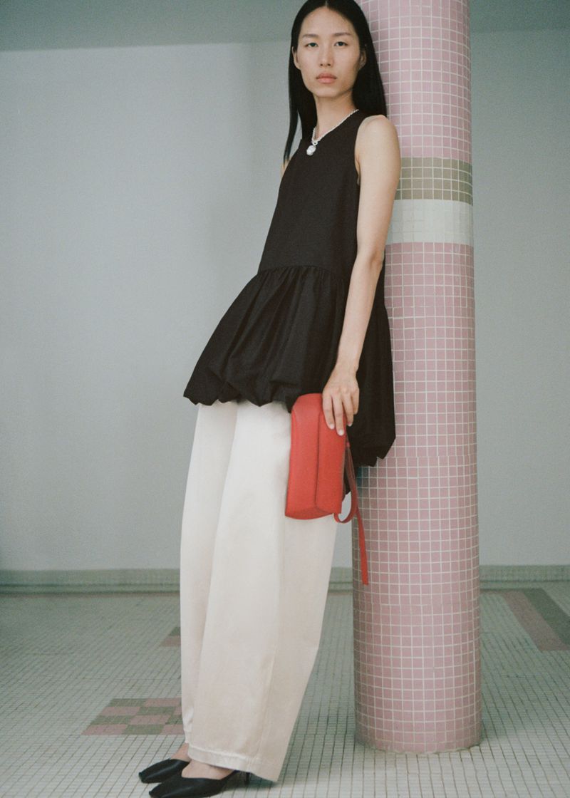 A stylish Asian woman in a black peplum top and white pants holds a red purse against a tiled column in a minimalist indoor setting.