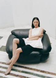 Model seated in a black leather armchair wearing a white sleeveless top and black shorts for Cordera Pre-Fall 25.