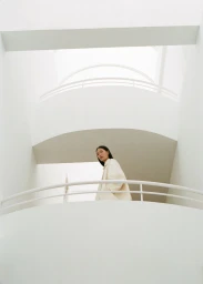 Model Yuanyuan Zhang in a cream jacket standing behind a white curved balcony inside a minimalist architectural atrium.
