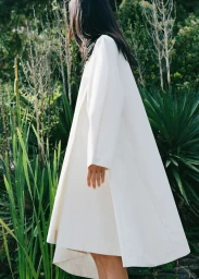 Side view of model Yuanyuan Zhang wearing an oversized white A-line coat surrounded by lush green foliage in an outdoor garden.