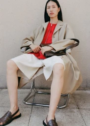 Model seated in a chrome chair wearing an open beige coat, red zip top, white shorts, and brown slingback loafers against a plain wall.