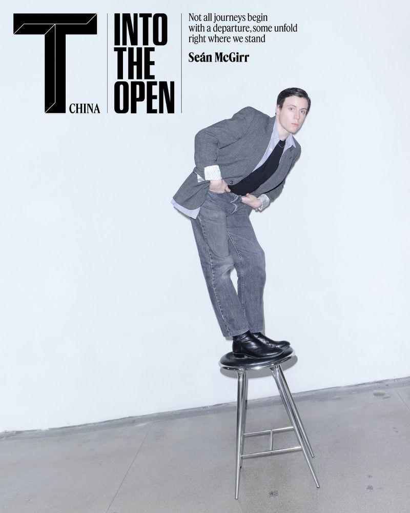 Seán McGirr standing on a metal stool wearing a grey blazer and jeans against a white studio wall for T Magazine China.