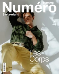 Antoni Porowski wearing a dark green shirt and yellow trousers reclining against a white wall for Numéro Homme Switzerland.