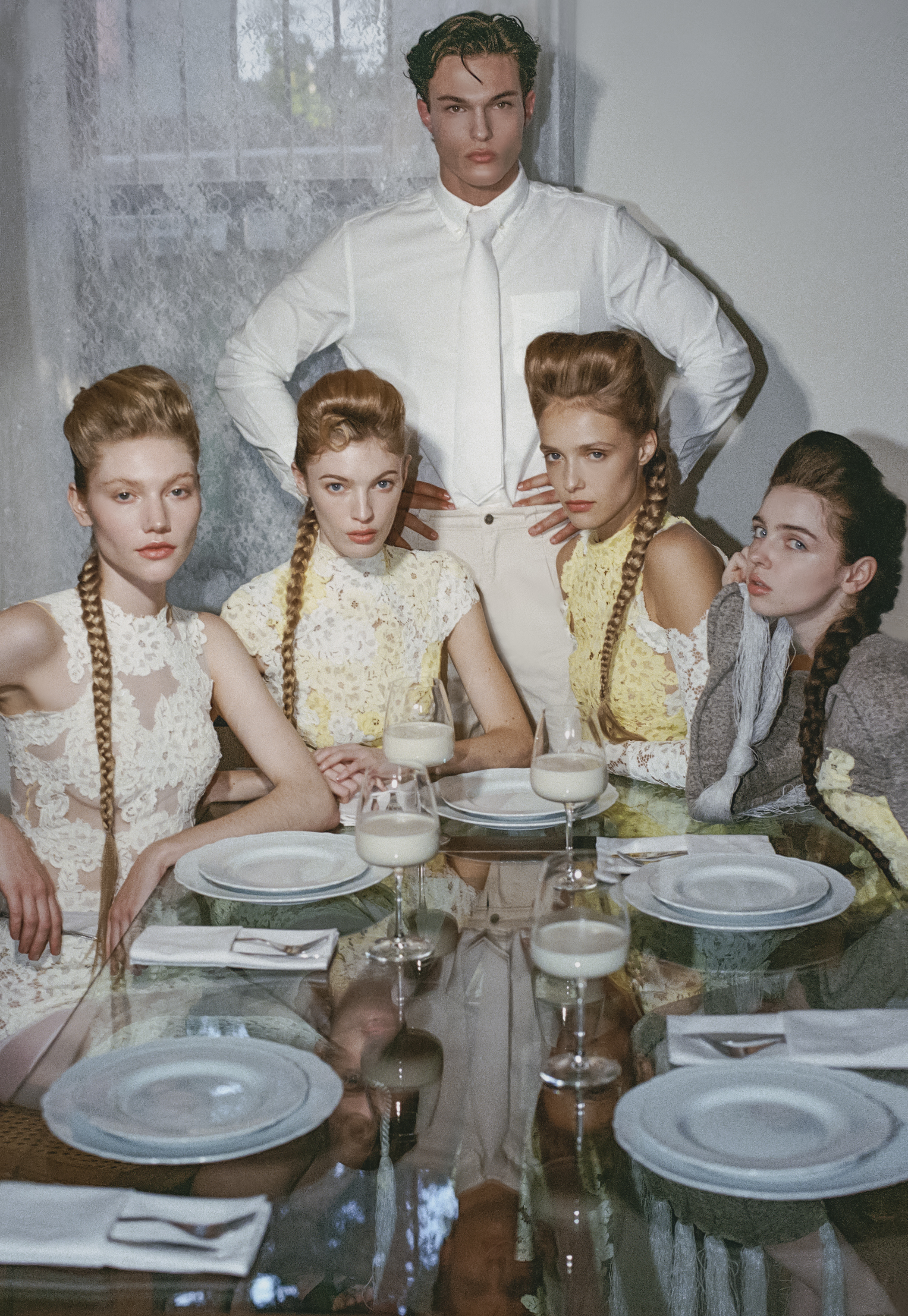 Four women with braided hairstyles seated at a glass dinner table with milk glasses while a man stands behind them.