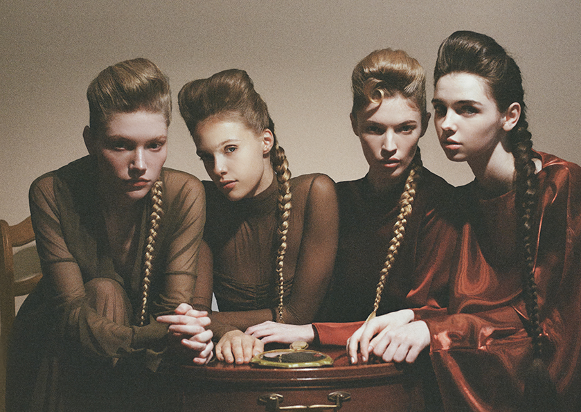 Four women with pompadour hairstyles and long braids pose closely around a wooden table wearing sheer brown tops and a rust satin dress.