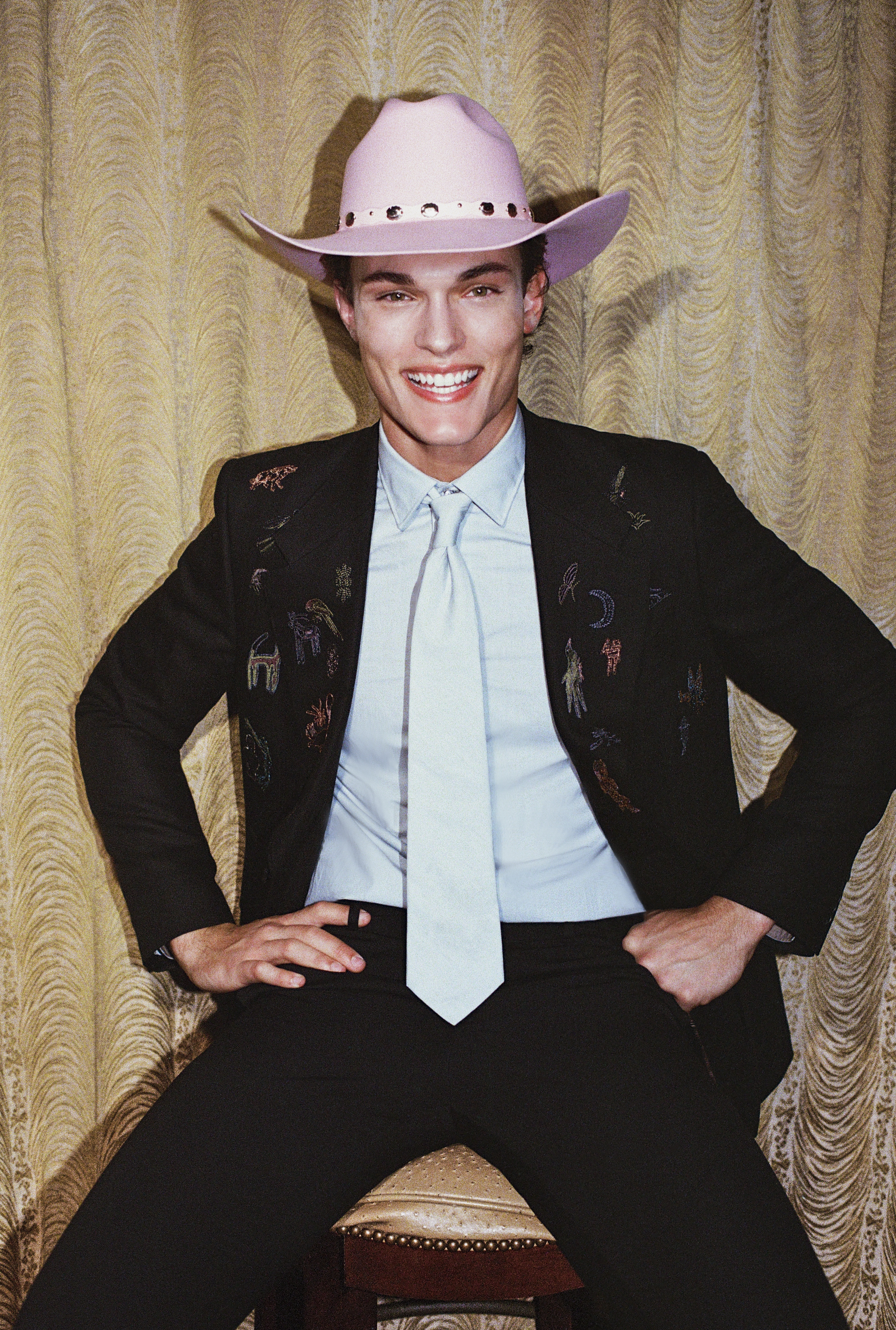 Smiling man in a pink cowboy hat, embroidered black suit, and light blue tie seated before textured gold drapes.
