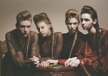 Four women with pompadour hairstyles and long braids pose closely around a wooden table wearing sheer brown tops and a rust satin dress.