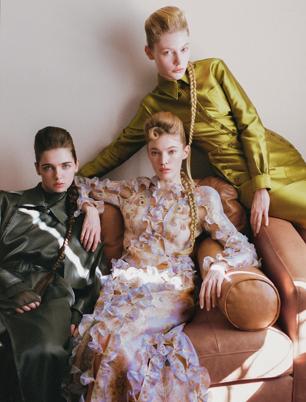 Three female models posing together, seated and leaning on a brown leather couch in an editorial fashion shoot by Zuzanna Blur.