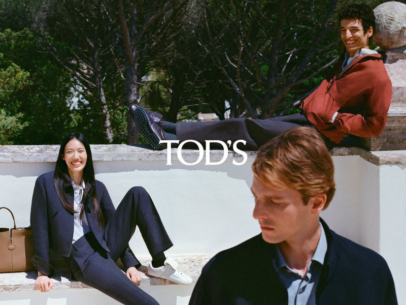 Three fashion models posing outdoors. The male models wear jackets, while the female model wears a blazer and pants with a tan tote.