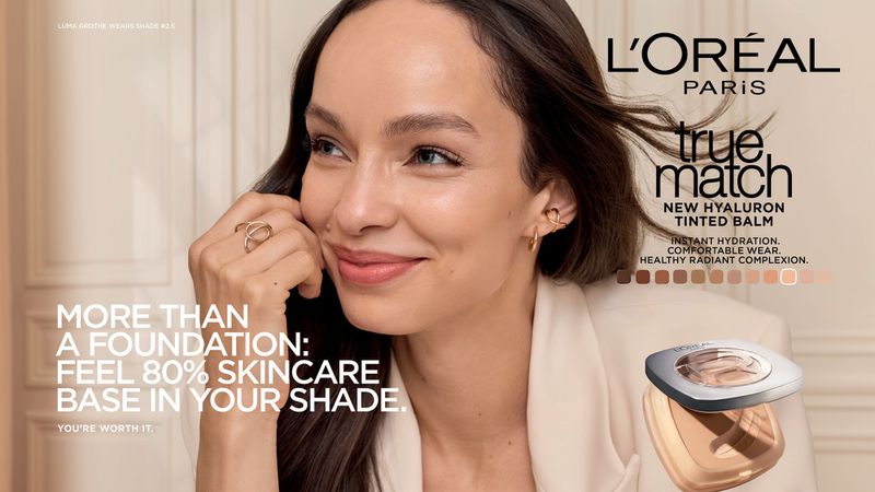 Model Luma Grothe smiling for the L'Oreal Paris True Match campaign in a white jacket against an interior wall.