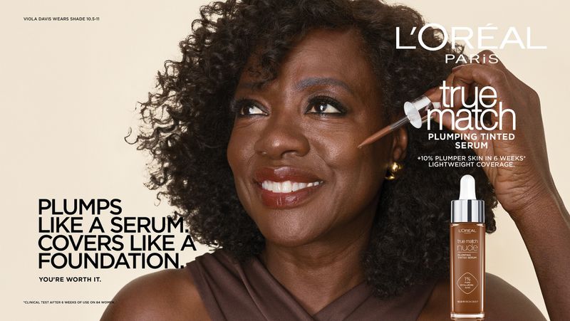 Viola Davis applying L'Oreal Paris True Match serum foundation with a dropper against a plain neutral background.