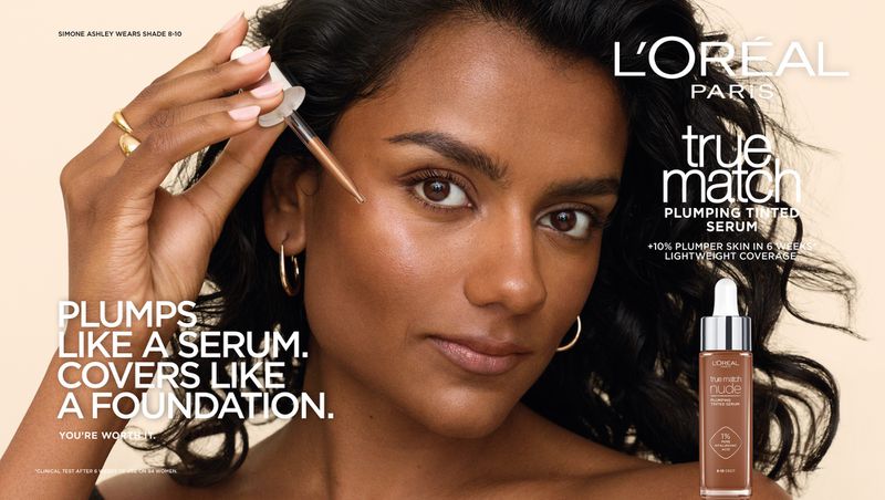 Simone Ashley applying tinted serum with a dropper for L'Oreal Paris True Match campaign against a warm neutral backdrop.