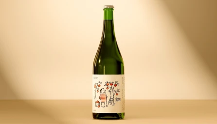 A green glass cider bottle with an illustrated apple orchard label and silver crown cap against a warm beige studio backdrop.