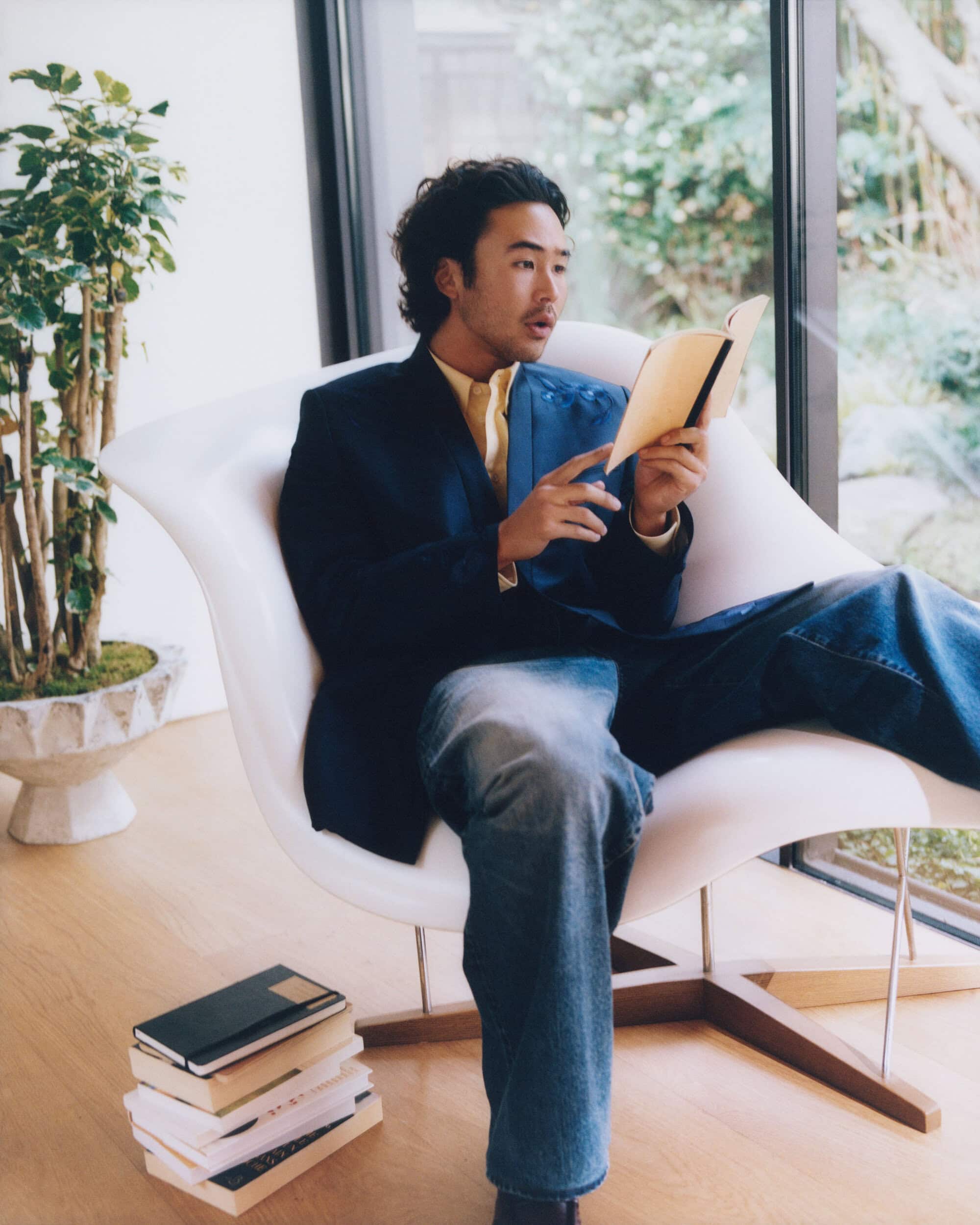 Nico Hiraga sits in a white modern lounge chair reading a book while wearing a navy blazer and wide-leg blue jeans.