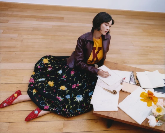 Model Lukita Maxwell seated on a wood floor in a brown leather jacket and floral embroidered black skirt sketching at a low table.