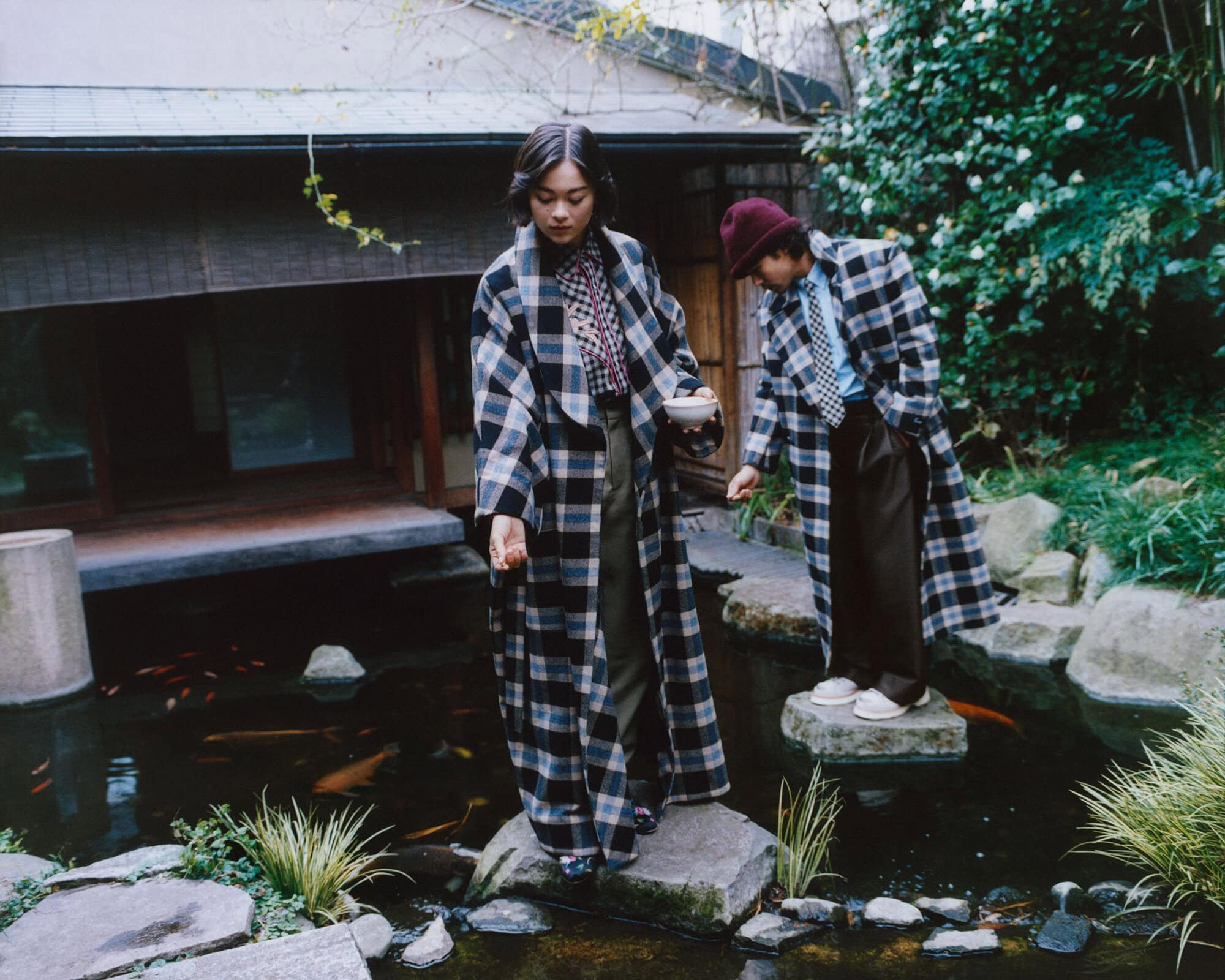 Lukita Maxwell and Nico Hiraga in oversized plaid coats standing on stepping stones over a koi pond in a Japanese garden.