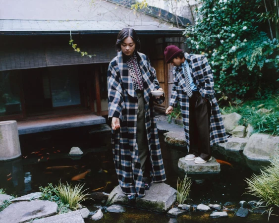 Lukita Maxwell and Nico Hiraga in oversized plaid coats standing on stepping stones over a koi pond in a Japanese garden.