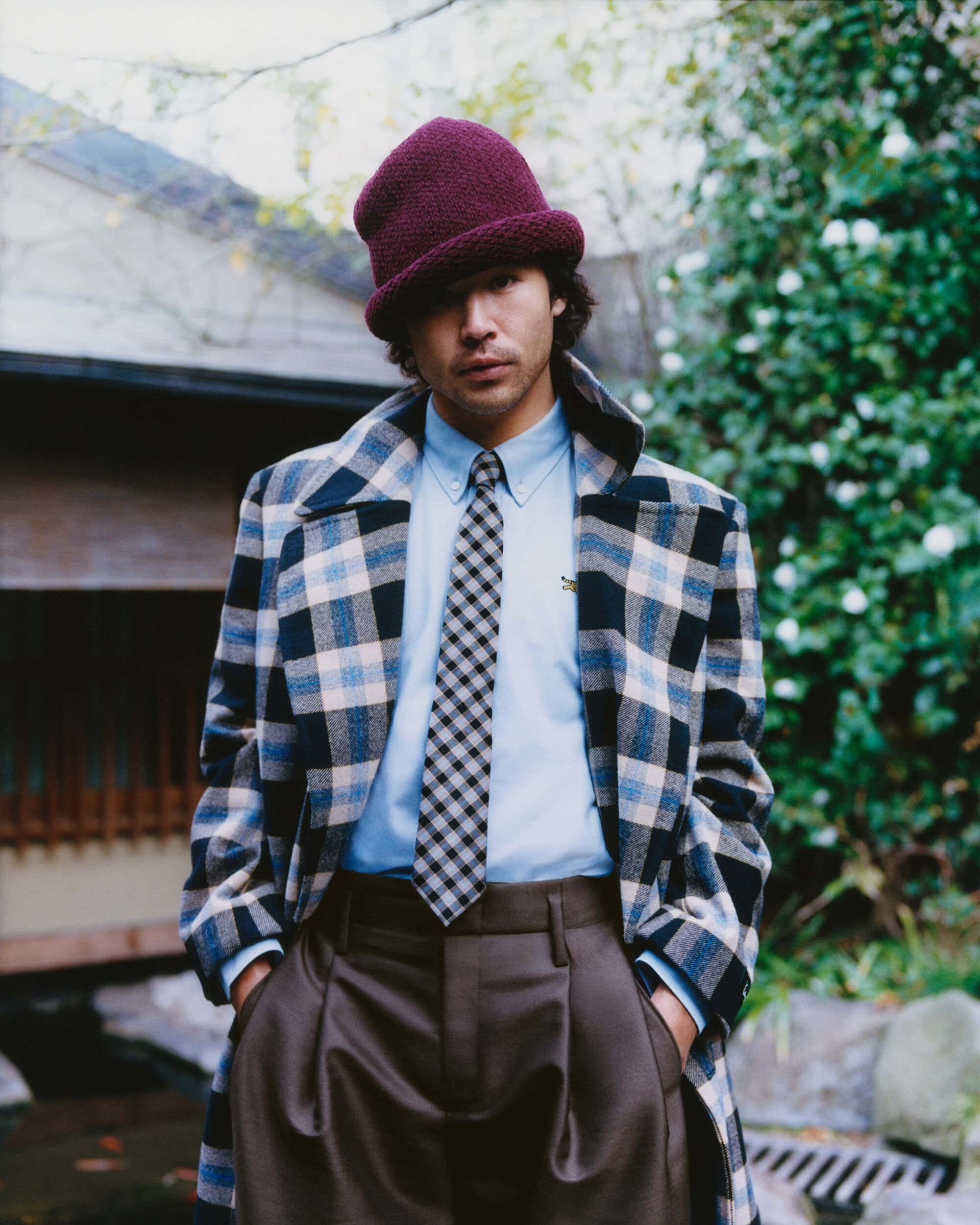 Man wearing a plum beanie, plaid coat, blue shirt, checkered tie, and brown trousers outdoors