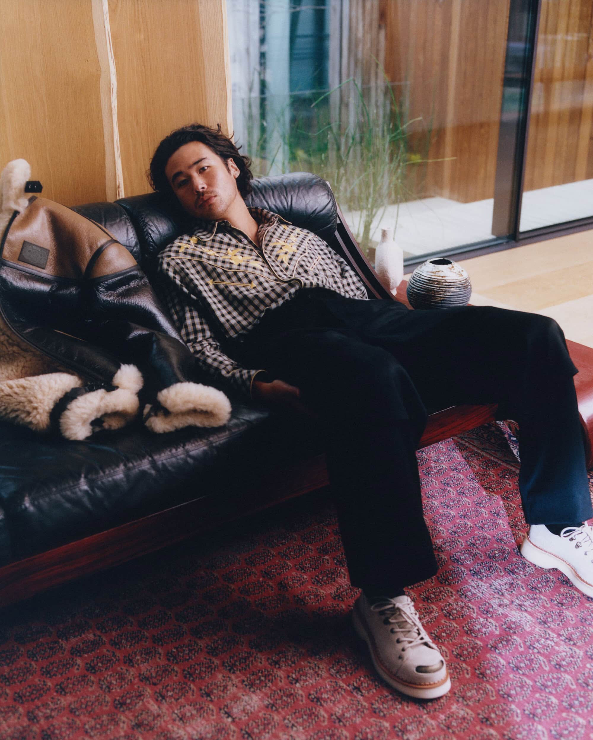 Nico Hiraga reclining on a black leather sofa wearing a plaid shirt, black trousers, and white sneakers for Kenzo.