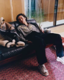 Nico Hiraga reclining on a black leather sofa wearing a plaid shirt, black trousers, and white sneakers for Kenzo.