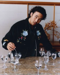 Nico Hiraga wearing a black floral embroidered jacket leans over a table filled with water glasses for Kenzo.