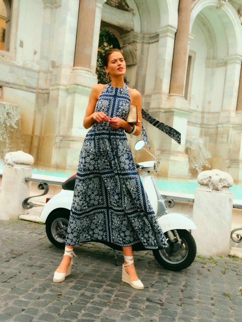 Model Bonnie Panasenko in a blue and white patterned halter maxi dress posing by a vintage white scooter near a stone fountain.