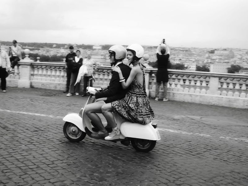 Two models riding a scooter together along a cobblestone scenic overlook in a black and white lifestyle photograph.