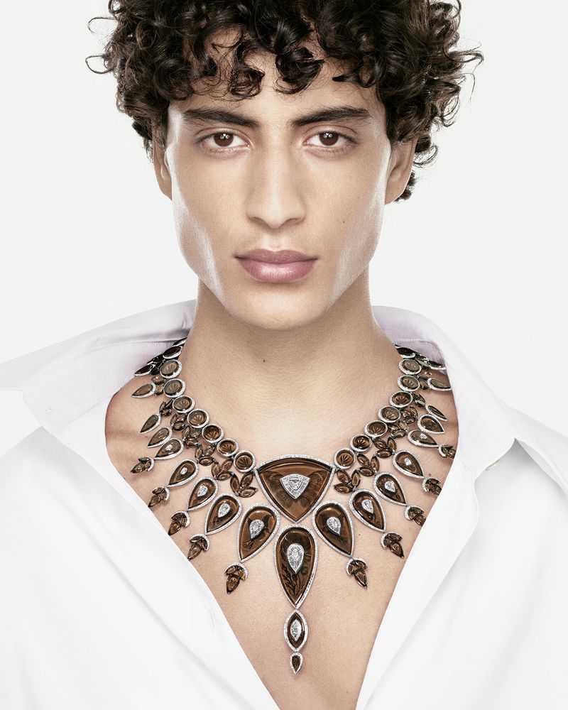 Model with curly dark hair wearing a large brown gemstone and diamond statement necklace with an open white shirt on white background.