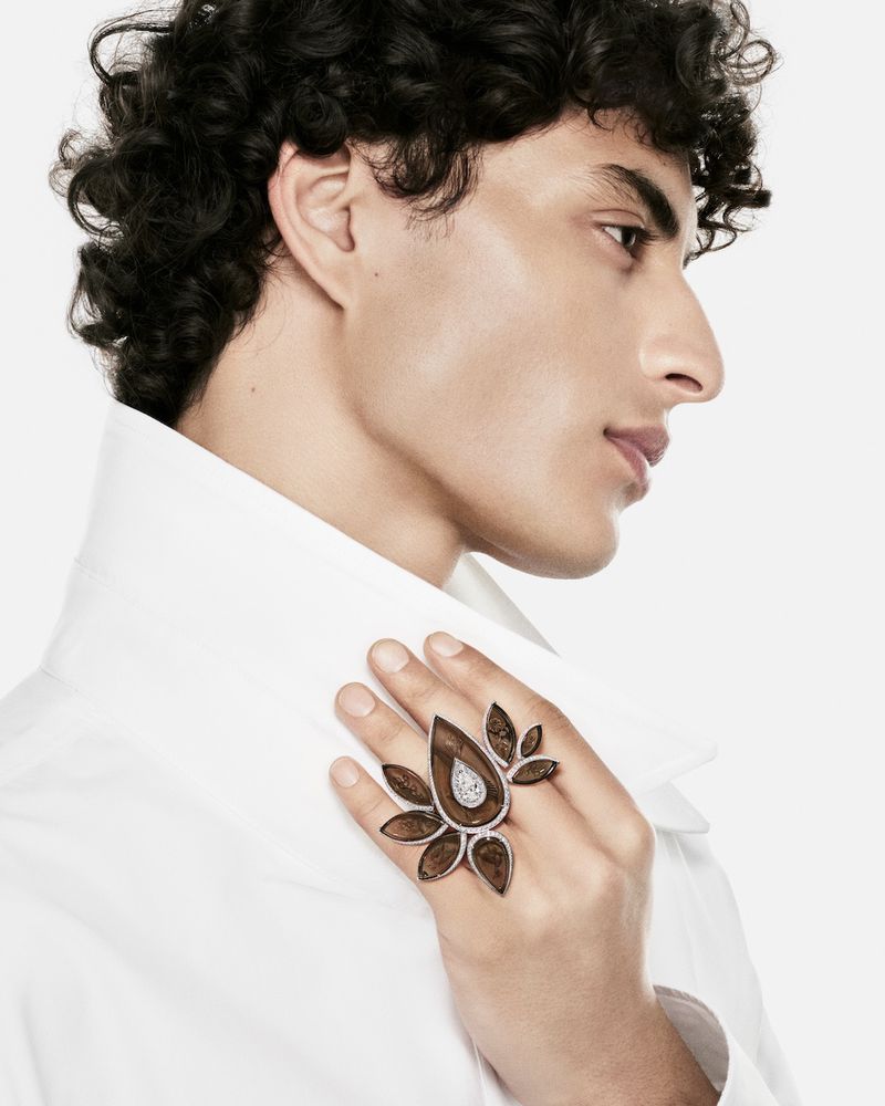 Male model in a crisp white shirt showcasing an elaborate floral high jewelry statement ring against a white background.